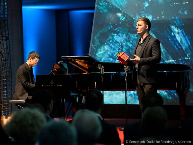 Ein schwarz gekleideter Pianist sitzt an einem Steinway Flügel und spielt, während rechts von ihm ein Sänger mit einem kleinen Geschenkpäckchen in der Hand steht und singt. Dahinter eine blaue Lichtprojektion.