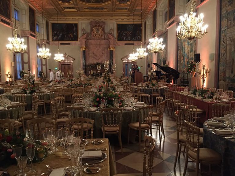 Blick auf den feierlich eingedeckten Kaisersaal der Residenz München. Links hinten prüfen zwei in weiß gekleidete Kellner die Tische. Rechts hinten steht ein Flügel. Davor zwei Musiker an ihren Instrumenten und spielen sich ein.