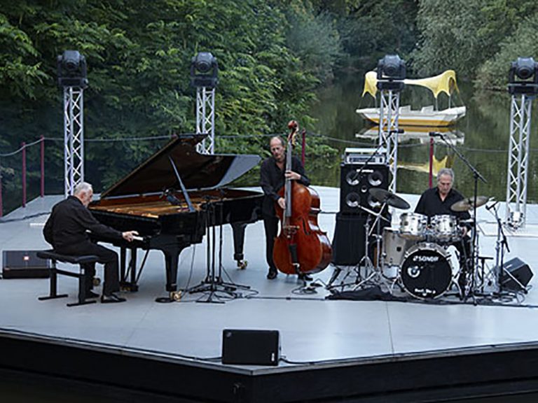 Auf einer Seebühne spielt links ein Pianist an einem Flügel. In der Mitte spielt ein Bassist seinen Kontrabass und rechts davon ein Schlagzeuger Schlagzeug. Alle drei sind schwarz gekleidet.
