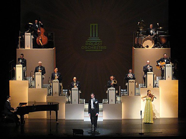 Musiker eines Orchesters im schwarzen Smoking auf einer Bühne mit ihren Instrumenten auf einzelnen Sockeln treppenförmig angeordnet. Im Vordergrund rechts eine Geigerin in einem gelb-glitzernden Kleid, in der Mitte ein Sänger im Frack, rechts ein Pianist an einem schwarzen Flügel.